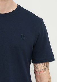 Άτομο που φοράει απλό μπλε ναυτικό T-shirt με μικρό λογότυπο ασπίδας στο στήθος και εμφανές τατουάζ με τριαντάφυλλο και ημερομηνία στο δεξί αντιβράχιο.