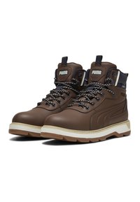 Botas de senderismo de cuero marrón de Puma con cordones negros y blancos, cuellos acolchados y suelas gruesas blancas con tacos marrones, logos laterales visibles.
