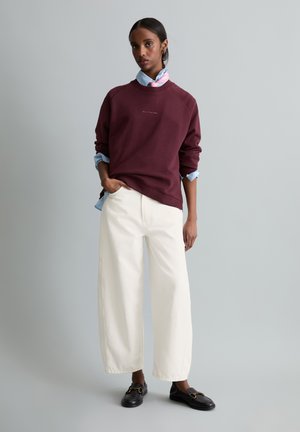 Femme debout avec une main dans la poche, portant un sweat-shirt bordeaux sur une chemise bleue, pantalon blanc large et mocassins noirs sur fond gris.