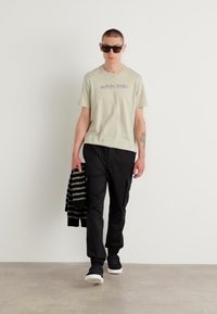 Lichtgroene T-shirt met tekst, zwarte cargobroek en zwarte sneakers. Gestreept zwart-wit jasje over de linkerarm gedrapeerd. Zonnebril op.