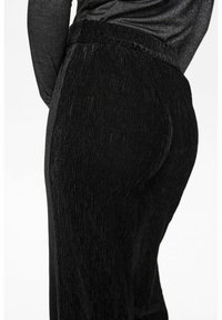 Zwarte corduroy broek met een gestructureerd oppervlak, voorzien van een elastische tailleband en een getailleerde pasvorm, zijkant gezien.