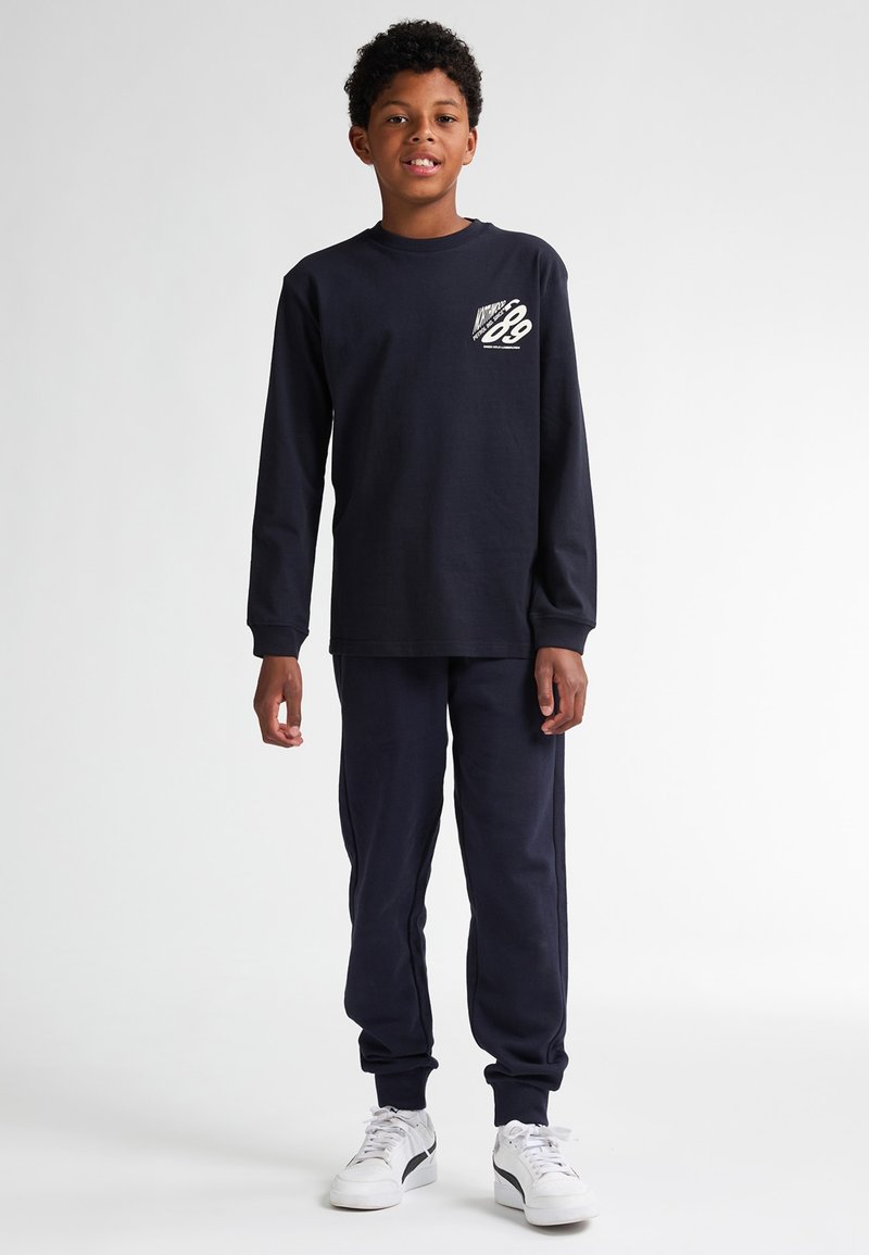 Maglietta a maniche lunghe blu navy con stampa grafica bianca sul petto, abbinata a pantaloni jogger coordinati e sneakers bianche. In cotone misto casual.