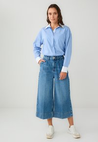 Vrouw staat met één hand in de zak, gekleed in een blauw gestreept shirt met een witte kraag en manchetten, brede cropped jeans en witte sneakers.