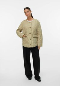 Quiltet beige jakke med knaplukning, to forlommer og en afslappet pasform. Båret over en hvid top og parret med sorte bukser.