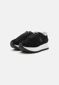 Sneaker in suede nera con intersuola bianca, suola esterna in gomma nera testurizzata, design sportivo con lacci e logo in rilievo sul lato.