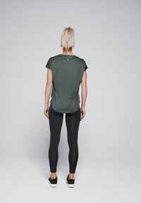 Donkergroene, lichte t-shirt met korte mouwen, gecombineerd met zwarte legging. Het model is van achteren gezien, wat een losse pasvorm laat zien.