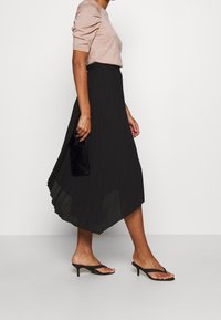 Zwarte geplooide midirok met een vloeiend ontwerp. Gecombineerd met een lichtroze nauwsluitend topje en zwarte sandalen met bandjes. Een clutch in de hand.