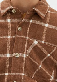 Brązowa koszula w kratę wykonana z miękkiego materiału, z zapięciem na guziki, dużym kołnierzem oraz kieszenią na piersi z białymi wzorami w kratkę.