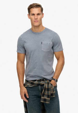 Gray cotton t-shirt with a round neckline, short sleeves, and a left chest pocket. Worn with dark jeans and a plaid shirt tied at the waist.