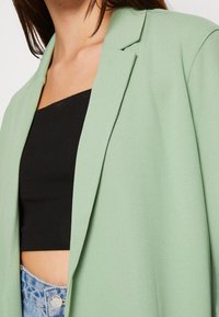 Ljusgrön kavaj med strukturerad design, slagkrage och en mjuk textur, bärs över en svart topp och denimshorts.