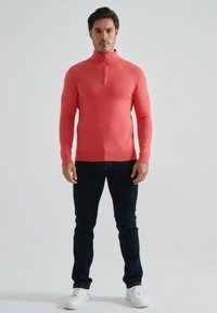 Pull-over corail en maille avec fermeture éclair à mi-hauteur, manches longues, coupe ajustée, porté avec un jean foncé et des baskets blanches, sur un fond uni.