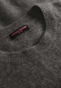 Grijze wollen trui met een ribbelkraag. Voorzien van een zwart label met "FRIEDA & FREDDIES" in roze tekst, met een zachte, fuzzy textuur.