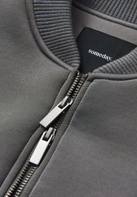 Giacca grigia con cerniera, collo a costine, cerniera in metallo e texture liscia. Etichetta del marchio "someday" visibile sul lato interno.