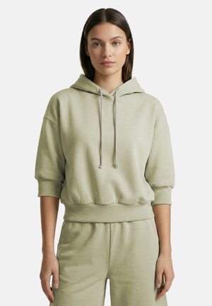 Junge Frau mit glatten braunen Haaren, die einen hellgrünen kurzen Hoodie mit Kordeln und passende Hosen trägt, steht vor einem weißen Hintergrund.