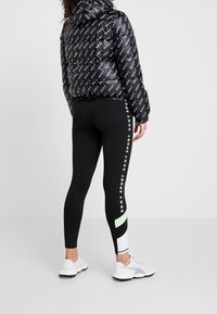 Černá péřová bunda s bílými logy, v kombinaci s černými legínami se světlým a mátově zeleným geometrickým vzorem a bílými sportovními botami.