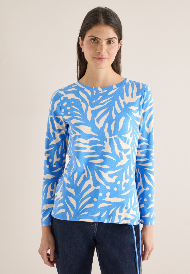 Femme portant un haut à manches longues bleu et blanc à motif de feuilles avec un col rond et un jean bleu foncé, debout devant un fond uni.