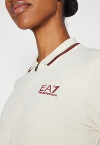 Detailní záběr osoby v krémovém polo tričku se zipem a burgunďským logem EA7 Emporio Armani na levé hrudi.