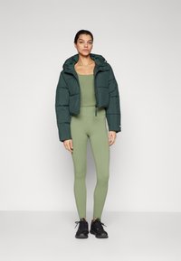 Veste courte en duvet verte avec capuche, assortie à des leggings moulants vert olive. Des chaussures de sport noires complètent la tenue.