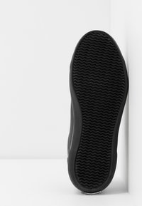 Semelle de sneaker noire avec un motif de grip texturé en zigzag. Le matériau semble caoutchouteux et flexible, avec un bord lisse.