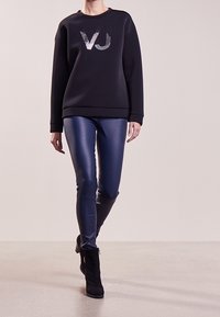Sweat-shirt noir avec un logo "VJ" argenté, associé à des leggings en simili cuir bleu brillant et des bottines noires sur un fond neutre.