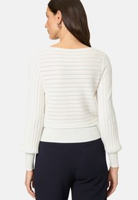 Cremefarbener Rippstrickpullover mit dezenten horizontalen Streifen, verkürzter Länge und enganliegenden Bündchen, kombiniert mit dunklen Hosen.