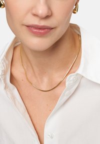 Goldfarbener Schlangenkettchen-Halsreif mit einem glatten, gebogenen Design, getragen über einem weißen Kragenhemd, mit einer glänzenden, reflektierenden Textur.