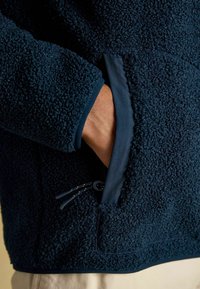 Käsi tummansinisen teksturoituneen fleece-takin vetoketjullisessa taskussa, näkyy hiha ja vaaleat housut.