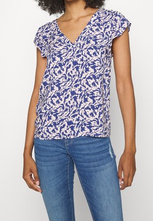 Femme portant un chemisier à manches courtes à motifs violets et blancs avec un col en V, assorti à un jean bleu, sur un fond blanc uni.