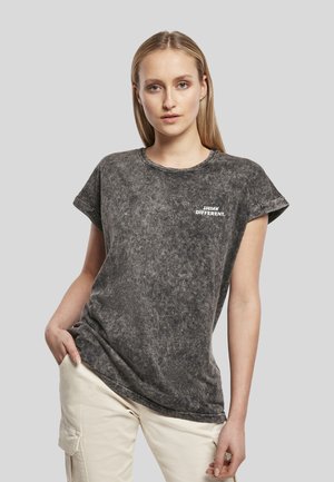 Jeune femme aux cheveux blonds raides portant un t-shirt noir délavé avec le texte "THINK DIFFERENT." et un pantalon beige clair sur fond gris.