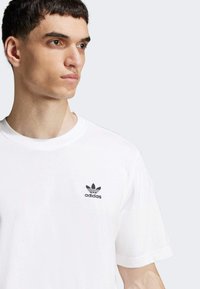 Hvid bomulds T-shirt med rund hals og korte ærmer. Har et lille sort Adidas-logo på venstre bryst. Glat tekstur.