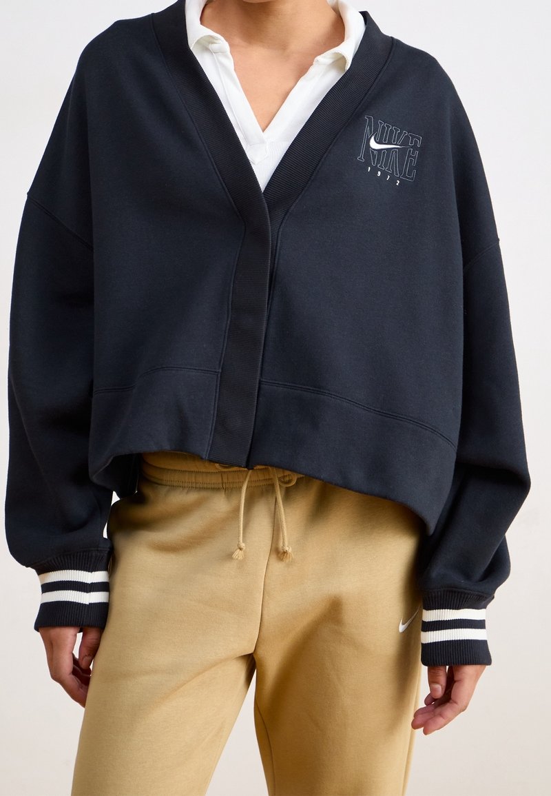 Sweatshirt cropped bleu marine avec un col en V, des poignets côtelés ornés de rayures blanches, et un logo Nike brodé sur la poitrine, associé à un pantalon de jogging beige.
