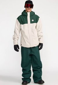 Heren snowboardoutfit met een beige en groene waterdichte jas met capuchon, gecombineerd met groene broek en zwarte handschoenen.
