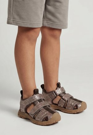 Dziecko ubrane w beżowe szorty i błyszczące brązowe sandały z zakrytymi palcami i podwójnymi paskami na rzepy, stojące na neutralnym tle.