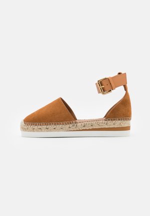 Brun ruskind espadrillesandal med ankelrem, guldfarvet spænde, mellemsål indpakket i jute og hvid gummiydersål mod hvid baggrund.