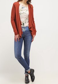 Rdzawoczerwony kardigan otwarty z lekkiego materiału, nałożony na kwiecistą bluzkę, w komplecie z dopasowanymi niebieskimi dżinsami i czarnymi botkami za kostkę.