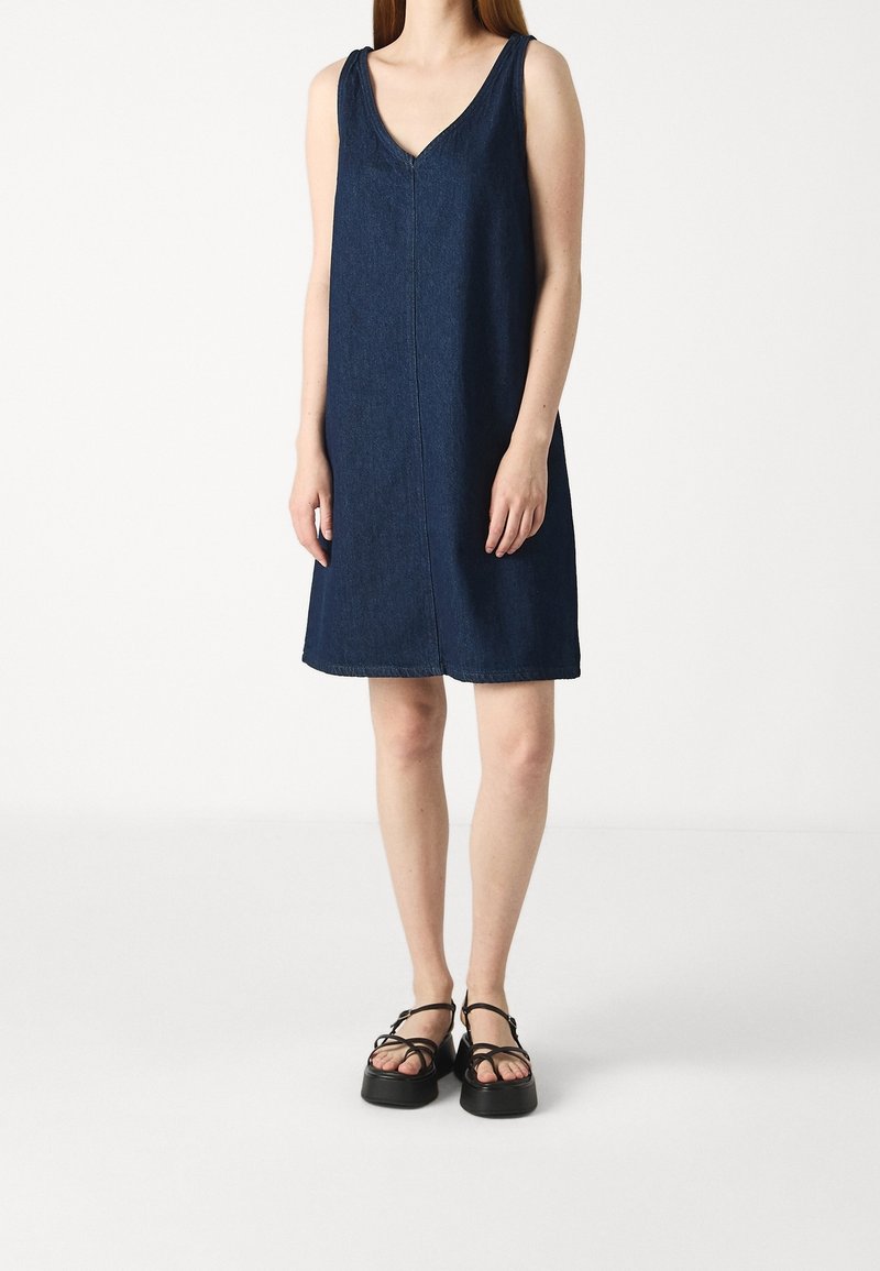 Femme portant une robe bleu foncé sans manches à col en V longueur genou et des sandales à plateforme noires, debout sur un fond blanc uni.