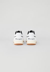 Vita sneakers med svarta accenter, med en texturerad gummisula och "Champion"-logotyp på hälen, visade bakifrån.