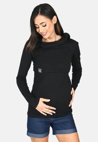 Top nero a maniche lunghe con scollo a cappuccio e texture a coste, abbinato a pantaloni corti in denim. Presenta un piccolo etichetta con logo sul davanti.