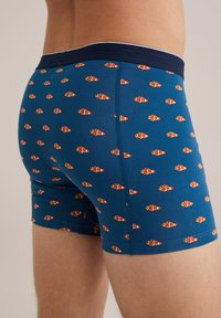 Boxersko perilo iz modre tkanine z motivom majhnih oranžnih klovnovskih ribic. Elastičen pas in gladka tekstura. Dolžina do sredine stegna.