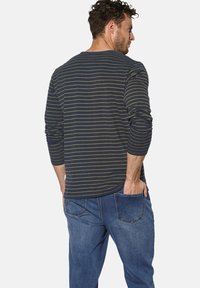 Mann mit Hand in der Gesäßtasche, trägt ein langärmliges Shirt in Marineblau und Olivgrün mit gestreiftem Muster sowie eine blaue Jeans, von hinten auf weißem Hintergrund.