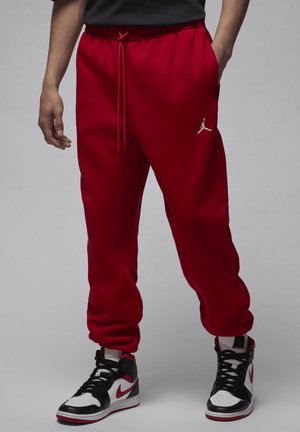 Homme portant un pantalon de jogging rouge avec cordon de serrage et des baskets montantes blanches, noires et rouges, debout sur un fond gris.