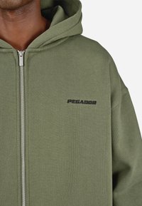Primo piano di una persona che indossa una felpa con zip di colore verde oliva, con la scritta "PEGADOR" ricamata in nero sul lato sinistro del petto.