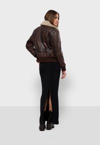 Veste en cuir marron de style bombardier avec un col en shearling beige, portée sur une jupe maxi noire avec une fente sur le côté, et des bottes noires épaisses.