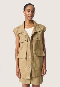 Khaki mouwloos vest met meerdere zakken, drukknoopsluiting en een hoge kraag. Gemaakt van stevig materiaal met een soepele textuur. Rechte snit.