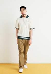 Beige polo skjorte med grønn krage og stripete kant, kombinert med khakibukser og hvite joggesko. Modellen står mot en ensfarget bakgrunn.