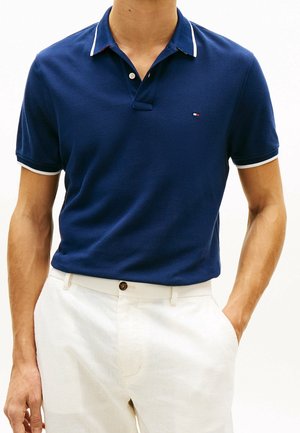 Homme portant un polo bleu marine à manches courtes avec bordure blanche et pantalon blanc, une main dans la poche, sur fond blanc.