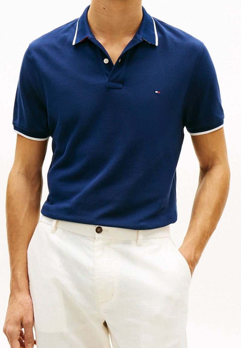 Homme portant un polo bleu marine à manches courtes avec bordure blanche et pantalon blanc, une main dans la poche, sur fond blanc.