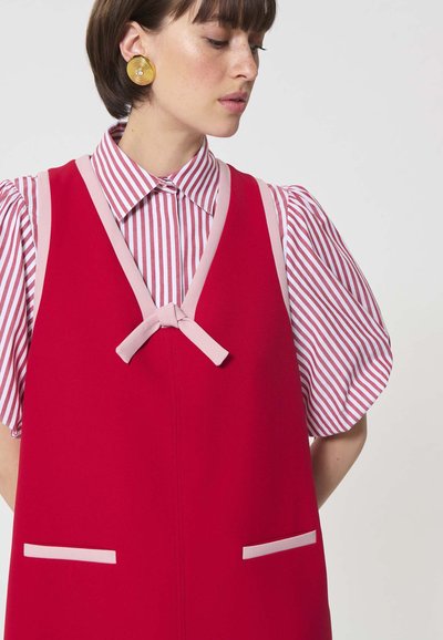 Femme portant des boucles d'oreilles circulaires dorées, une robe rouge sans manches avec un bord rose, par-dessus un chemisier à manches bouffantes rayé rose et blanc, regardant vers le bas.