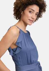 Robe bleue sans manches avec détails à volants sur les épaules, corsage ajusté et taille nouée. Confectionnée en tissu lisse avec des plis subtils.