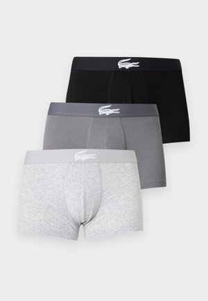 Atletický tmavší muž stojaci bosý, oblečený v čiernych boxerkách s malým bielym logom na páse na jednoliatom bielom pozadí.
